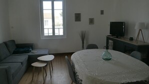A living room with a sofa, two small tables, a TV, and a window with blinds. - L'océane du pays - Saint hilaire de Riez (SAINT HILAIRE DE RIEZ)