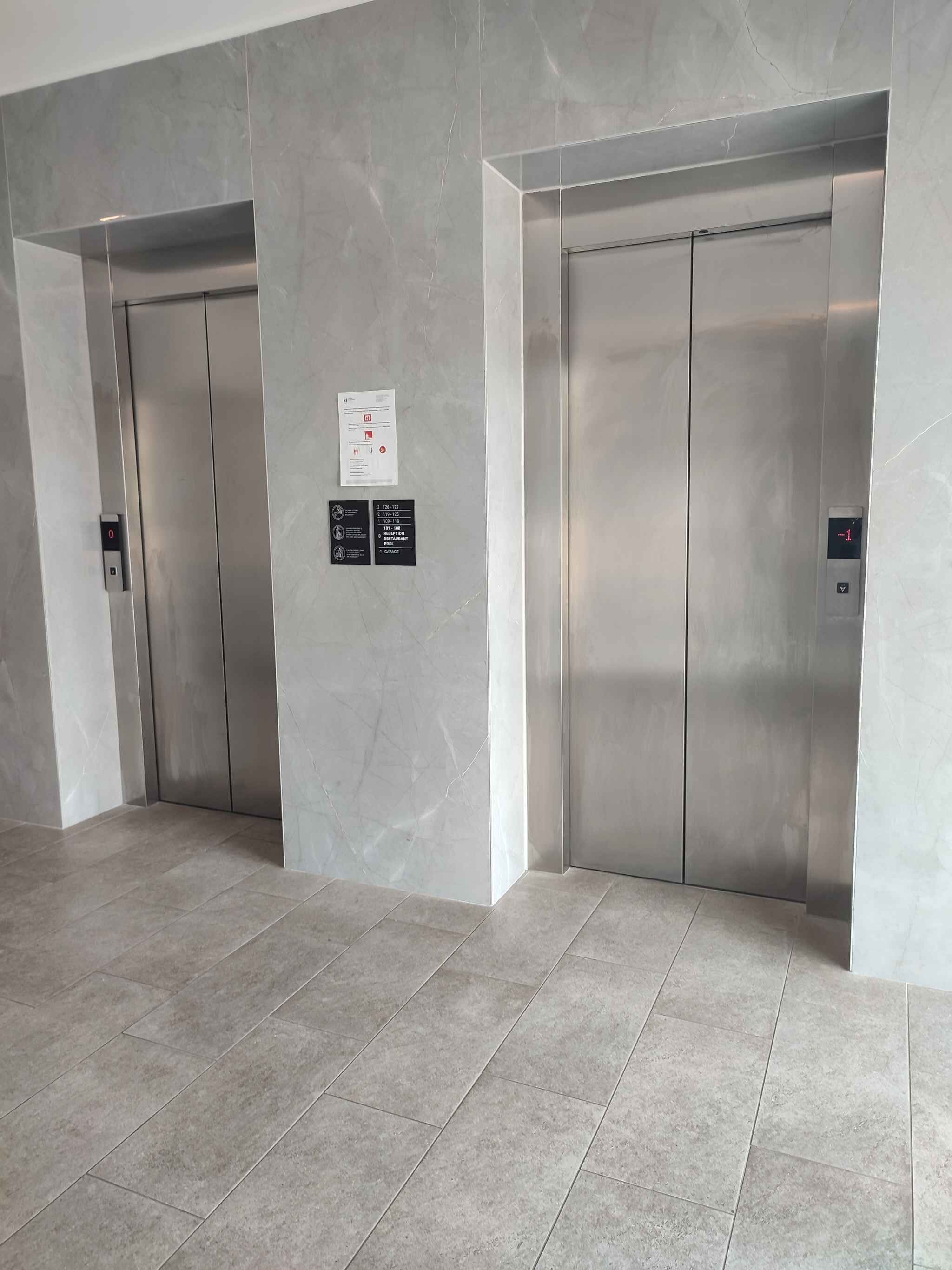 To elevatordøre i en moderne bygning med et marmorgulv og et skilt, der viser vej til restaurant og parkeringskælder.