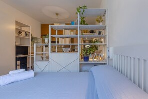 A bedroom with a bed, a white radiator, a white shelf unit with books and plants, and a television on a stand. - GuestHost - Bright studio apartment perfect for two people, located in a building with a garden in the Cirenaica district, an area near the center equipped with all services. Located just 20 meters from the entrance to the S.Orsola Polyclinic, it is idea (Bologna)
