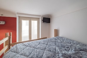 Une pièce comprenant un lit, un téléviseur fixé au mur, une fenêtre avec des stores et une porte.