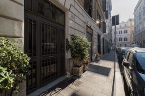 Een smalle stadstraat met geparkeerde auto's, een gebouw met een grote metalen poort, en potplanten langs de stoep.