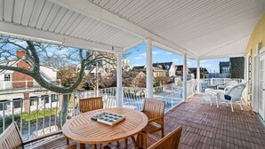 A covered outdoor patio with a round wooden table and chairs, white railings, and a view of surrounding buildings.