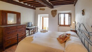 A bedroom with a wooden dresser, a desk with a computer, a bed with pillows, and a stone wall. - Stunning apartment in Monticchiello with WiFi (Monticchiello)