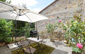 En udendørs terrasse med bord og stole, grill og en bygning i sten i baggrunden.