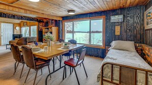 A cozy cabin interior with a wooden ceiling, a dining area with a table and chairs, a bed with a metal frame, and a large window offering a view of a lake.