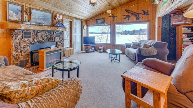 A cozy cabin interior with a stone fireplace, wooden walls, a television, and a large window overlooking a snowy landscape.