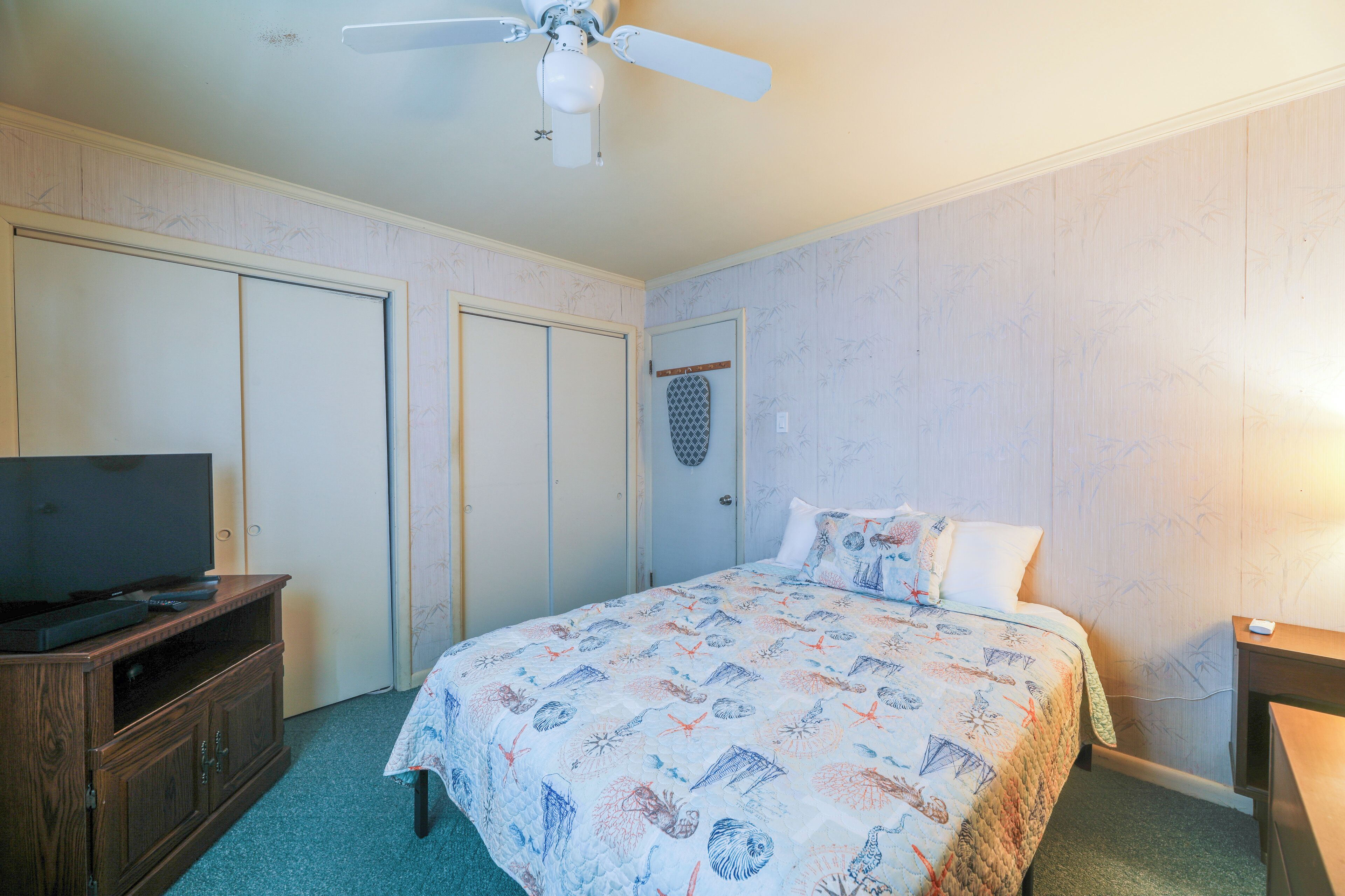 Une chambre à coucher avec un lit, un téléviseur posé sur un meuble en bois, un ventilateur de plafond et un luminaire fixé au mur.
