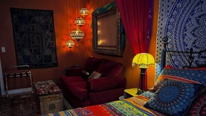 Une chambre avec un grand lit, un fauteuil rouge, une petite table et une tapisserie murale.