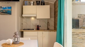 A compact kitchen with a gas stove, a sink, and cabinets. There is a small table with a lantern and a decorative item. A picture hangs on the wall, and a window with a view of the outside is visible. - Sea Sound Loft Naxos (Giardini Naxos)