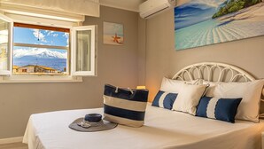 A bedroom with a bed, a window with a view of a mountain, an air conditioner, and a painting of a beach. - Sea Sound Loft Naxos (Giardini Naxos)