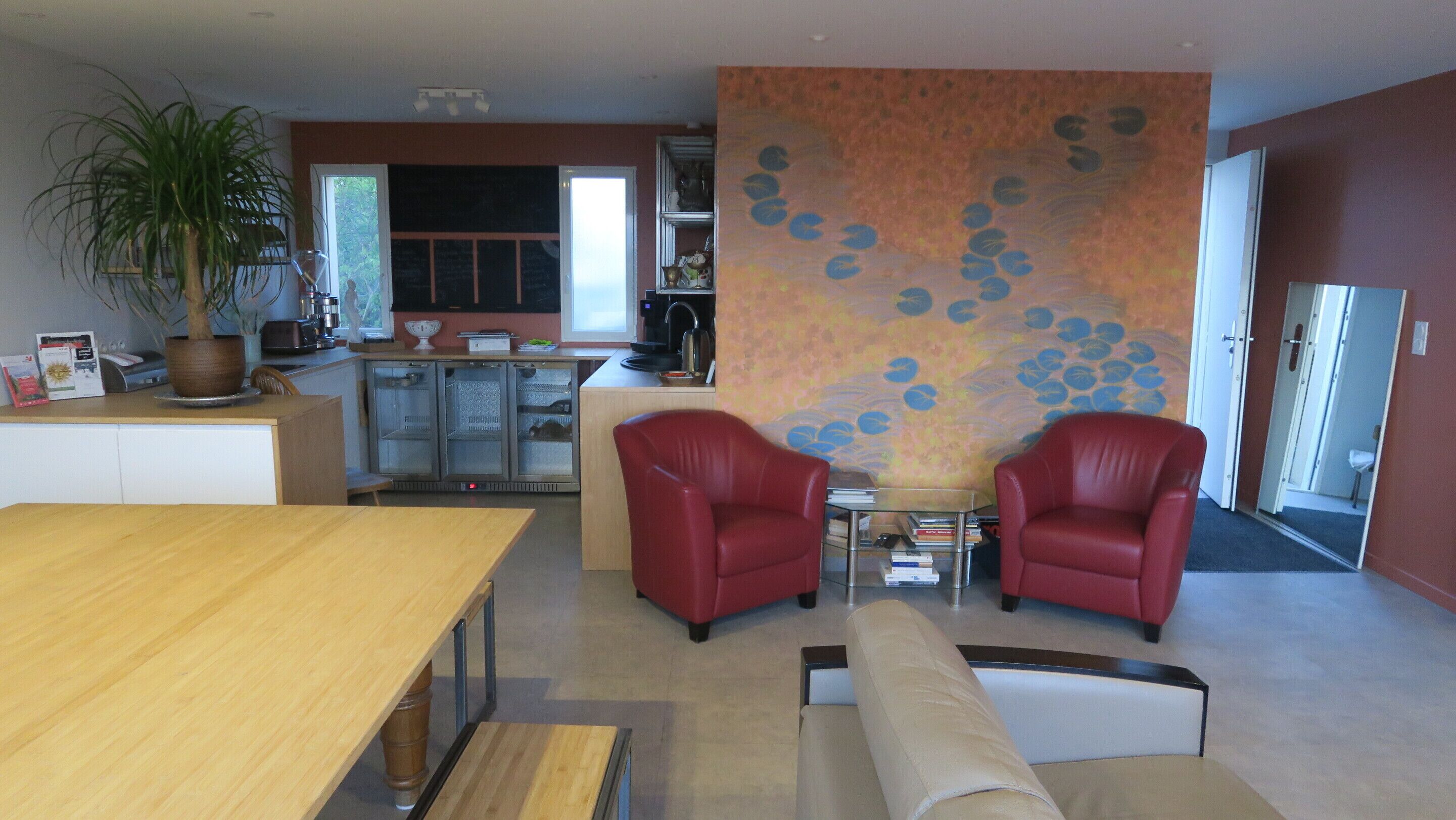 A modern living room with a wooden dining table, two red armchairs, a beige sofa, and a large potted plant.