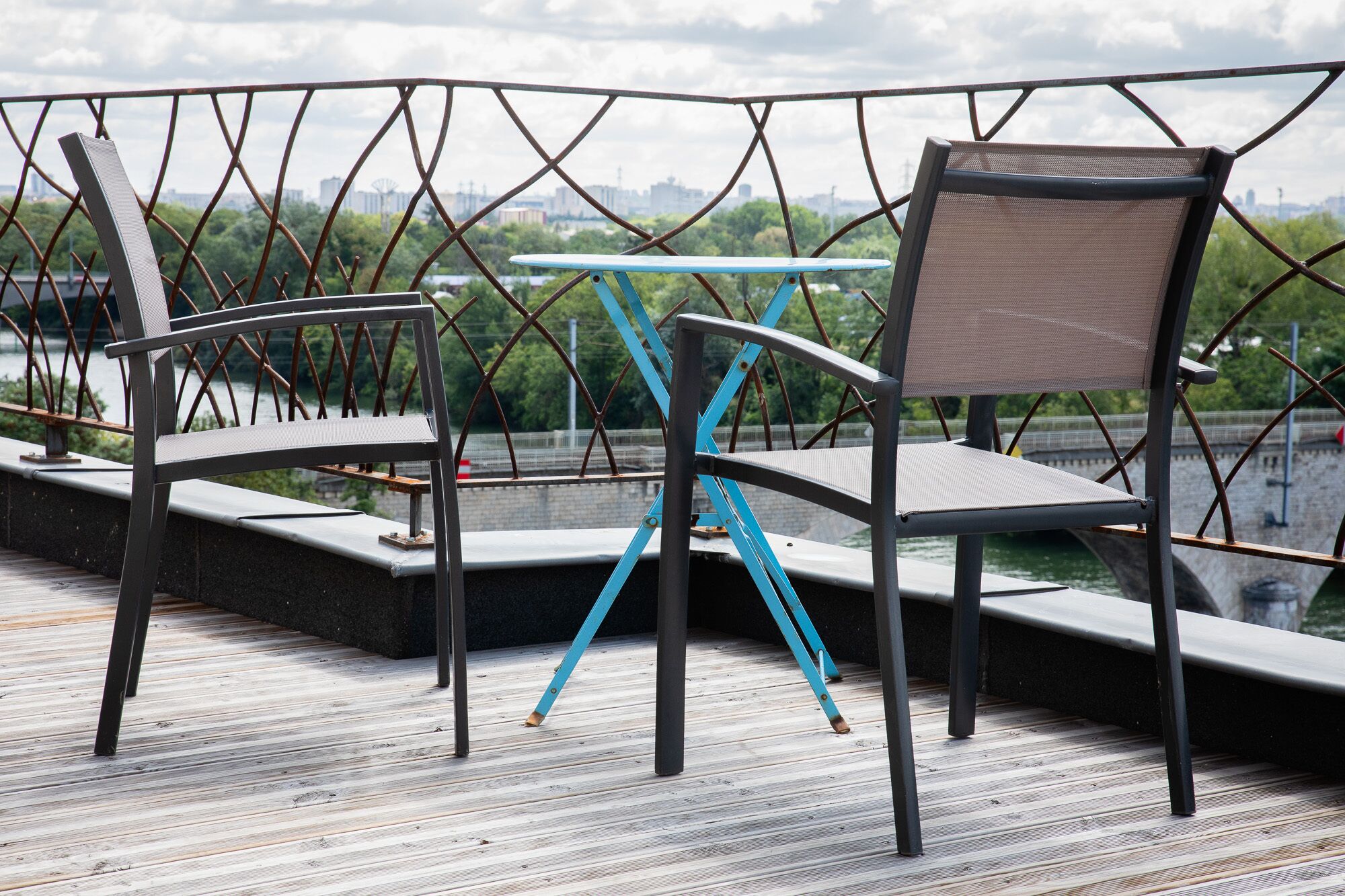 A deck with two chairs and a table, overlooking a river with a cityscape in the background.