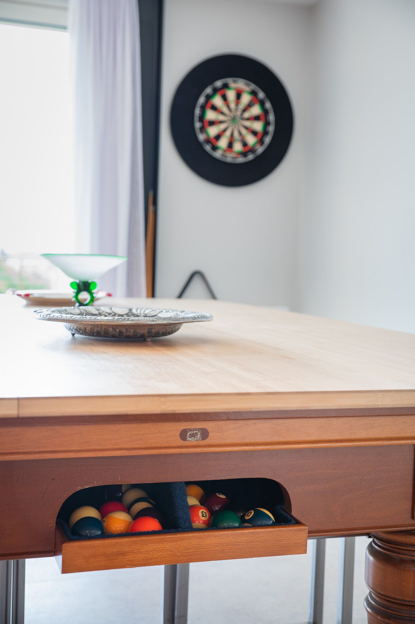 A dartboard mounted on a wall, a dartboard on a table, and a drawer with billiard balls.