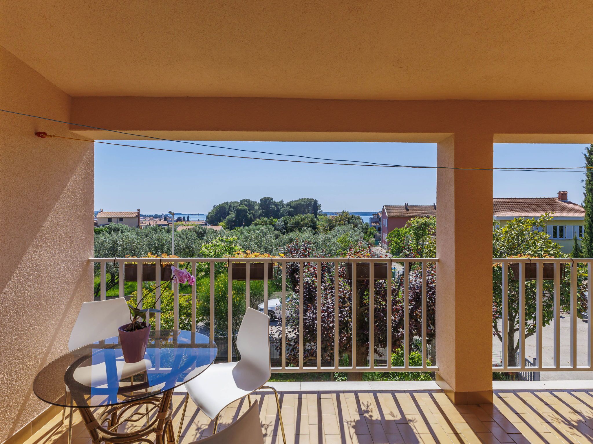 Un balcon avec une table et des chaises en verre, une plante en pot et une vue sur un paysage côtier, avec des arbres et des bâtiments.