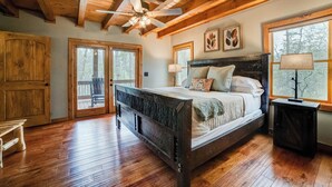 A spacious bedroom with a large bed, wooden ceiling beams, and a view of the outdoors through a window.