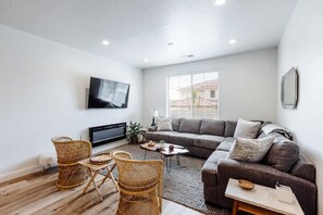 A modern living room with a flat-screen TV, a grey sofa, wicker chairs, and a coffee table.