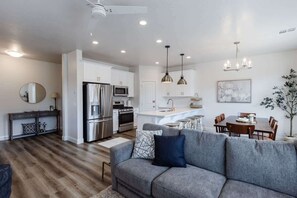 A modern kitchen with stainless steel appliances, a dining area with a wooden table and chairs, and a living room with a grey sofa. - Desert Riviera | Prime Location For Adventure! (Washington)