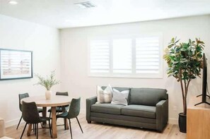 A modern living room with a grey sofa, a round dining table with chairs, a potted plant, and a window with blinds.