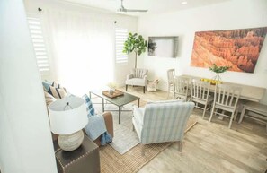 A living room with a sofa, armchair, coffee table, and a dining set. There is a plant and a painting on the wall.