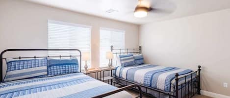 Une chambre avec deux lits, chacun doté d’un cadre en métal et de draps bleus. Il y a deux tables de chevet avec des lampes, et la chambre est équipée d’un ventilateur de plafond.