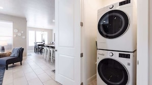 A laundry room with two stacked front-loading washing machines, a dining area with a table and chairs, and a door leading to another room.