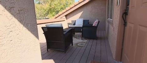 Een overdekt terras buiten met een tafel, stoelen, een deur en uitzicht op bomen.