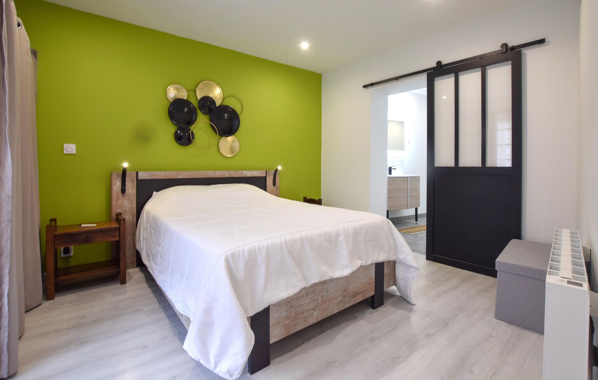 A bedroom with a bed, a nightstand, a wall-mounted light, a sliding door, and a small trash bin.