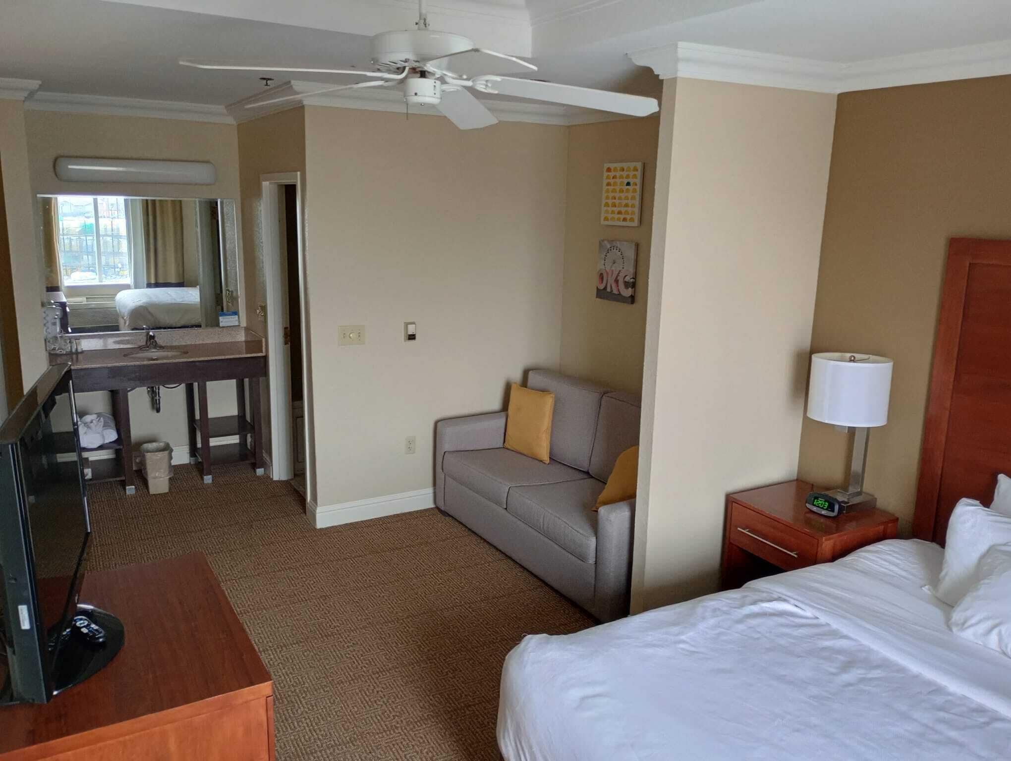 A hotel room with a bed, a desk, a sofa, a TV, a lamp, and a ceiling fan.