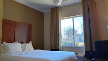 A hotel room with a large bed, a wooden headboard, a ceiling fan, a window with curtains, and a chair.