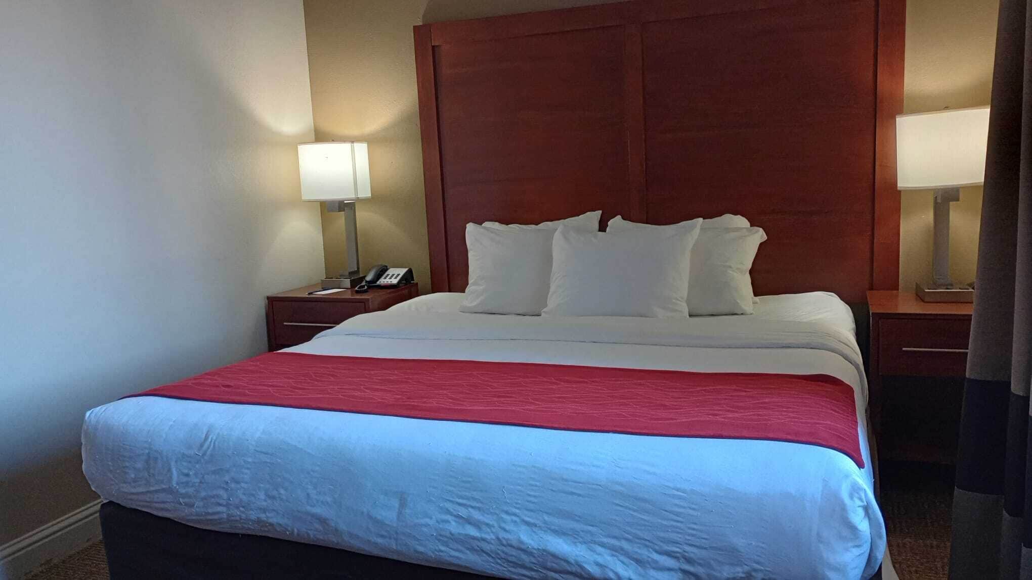 Une chambre d’hôtel avec un grand lit, une tête de lit en bois, deux tables de chevet avec des lampes et une touche de couleur rouge sur la couvre-lit.