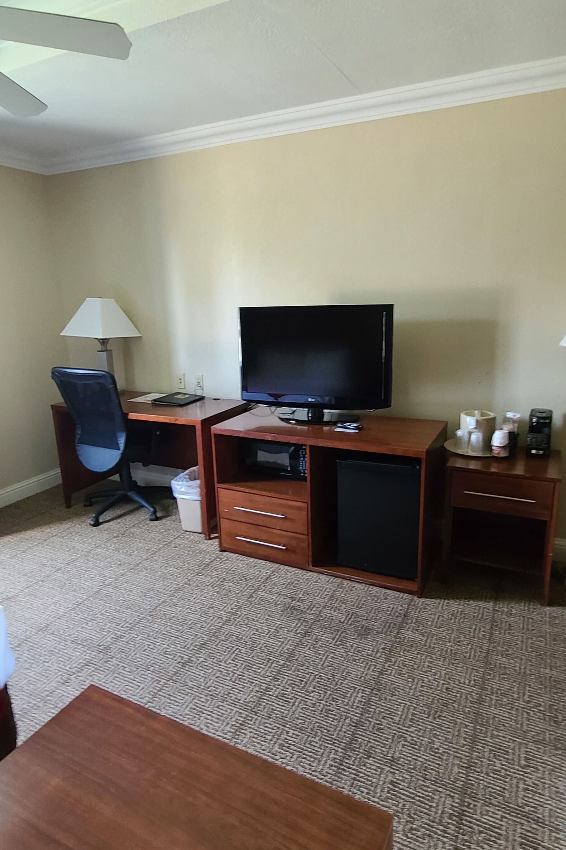 A hotel room with a wooden desk, a television, a lamp, a trash bin, and a coffee machine.
