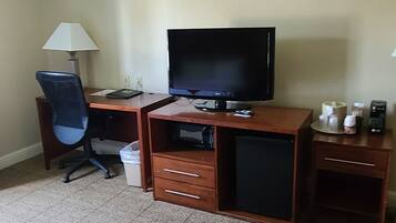 A hotel room with a wooden desk, a television, a lamp, a trash bin, and a coffee machine.