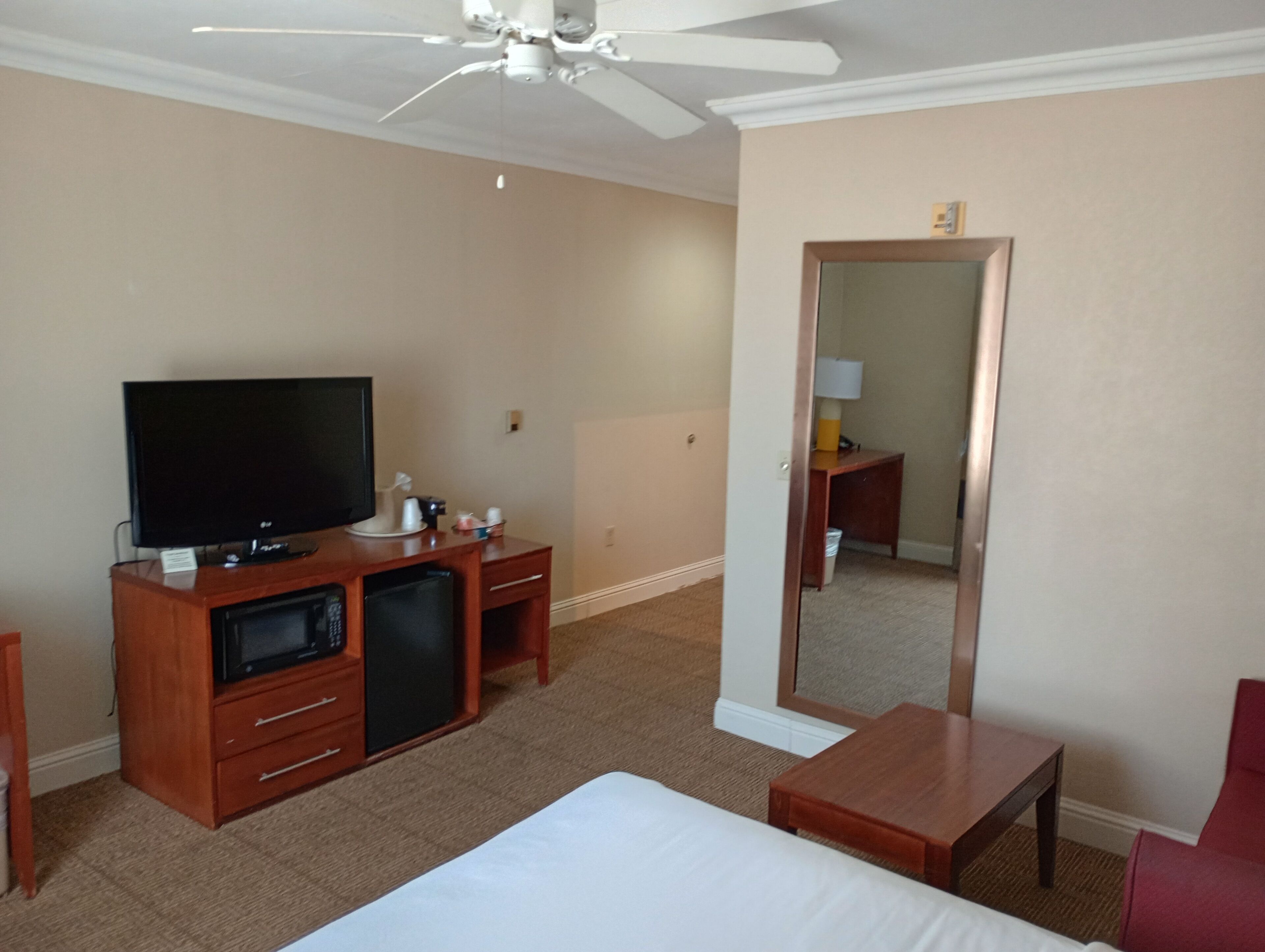 A hotel room with a bed, a TV, a desk, a chair, a mirror, and a ceiling fan.