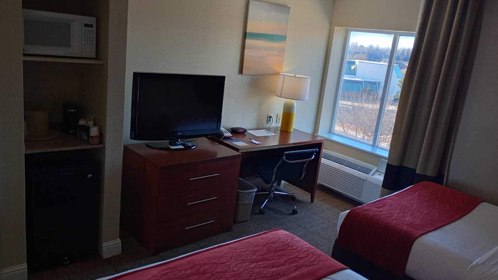 Et hotelværelse med en seng, et skrivebord med en computer, et fjernsyn og et vindue med udsigt til bygninger.