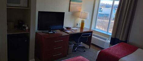 A hotel room with a bed, a desk with a computer, a TV, and a window with a view of buildings.