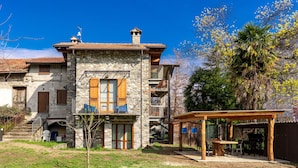 A stone building with a balcony, a wooden pergola, and a grassy yard.