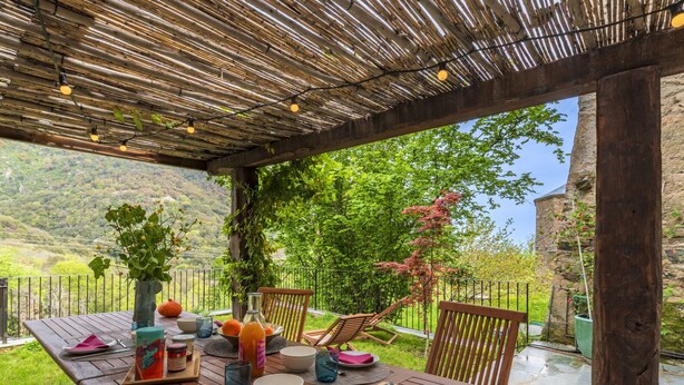 Een terras met een houten tafel voor twee personen, omringd door stoelen, met uitzicht op een weelderig landschap en een stenen gebouw op de achtergrond.