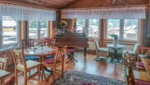 Ein gemütliches Zimmer mit Holzwänden und -decken, einem Klavier, einem Esstisch mit Stühlen und großen Fenstern mit Spitzenvorhängen.
