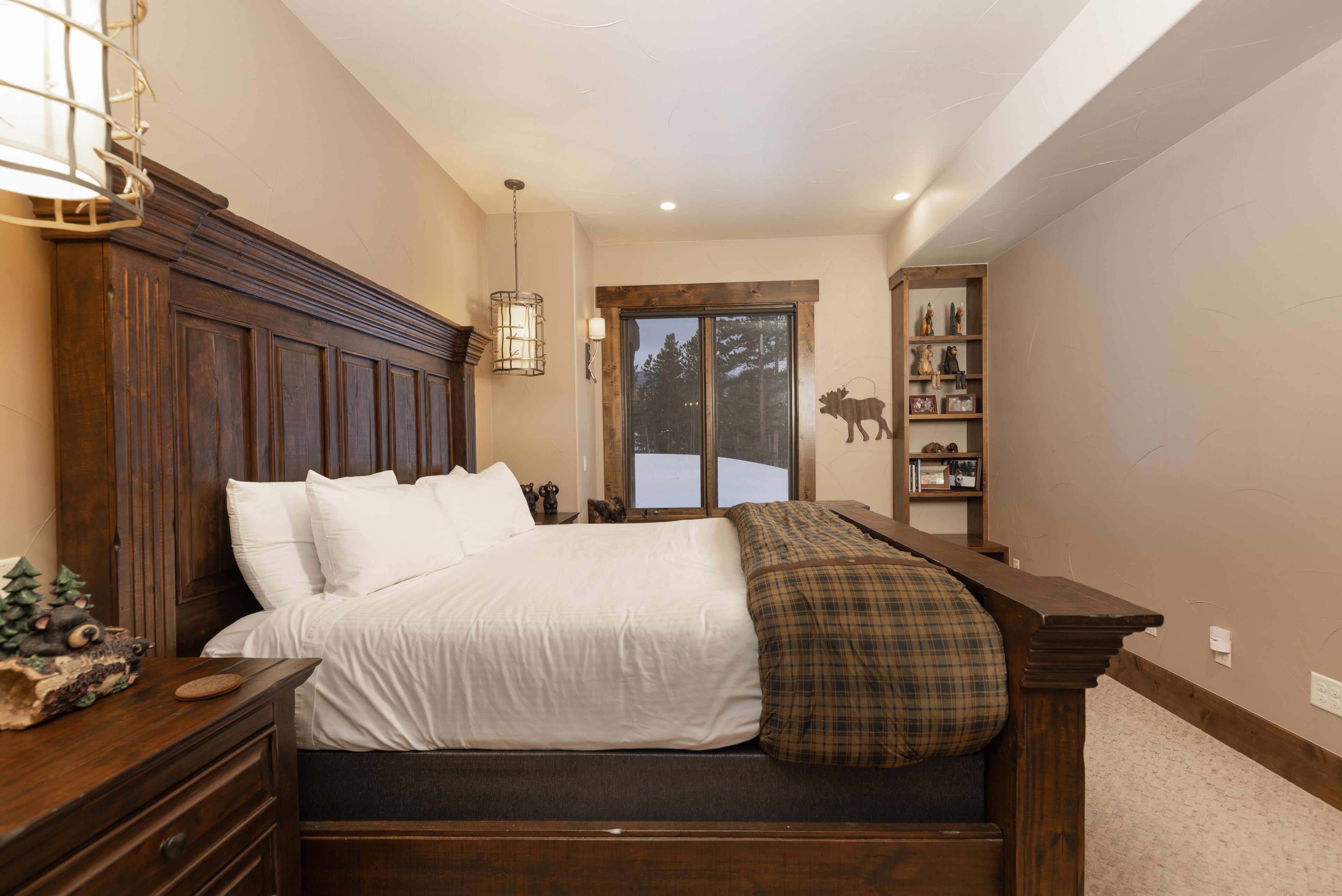 A bedroom with a large bed, wooden headboard, a bedside table with a lamp, a shelf with decorative items, and a view of the outdoors through a window.