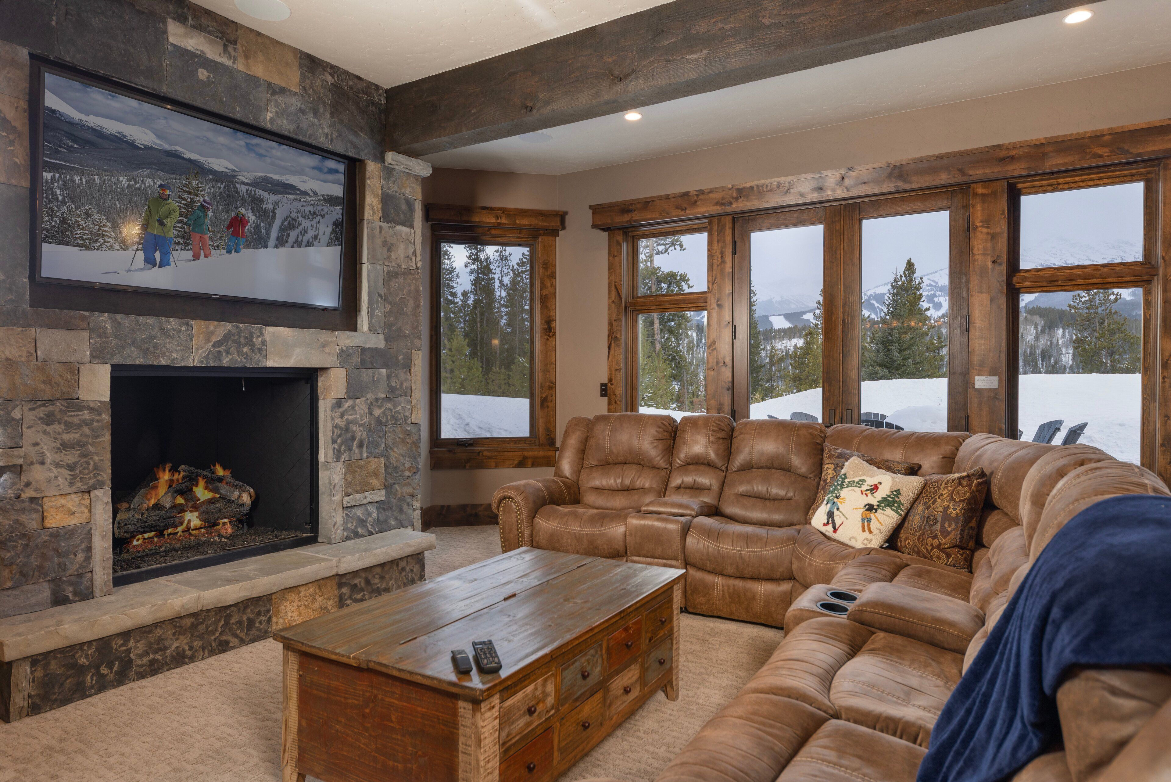 A cozy living room with a stone fireplace, leather recliner, and large windows offering a snowy mountain view.