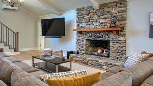 Una sala de estar con chimenea de piedra, un televisor de pantalla plana montado en la pared, una escalera de madera y un sofá seccional grande.