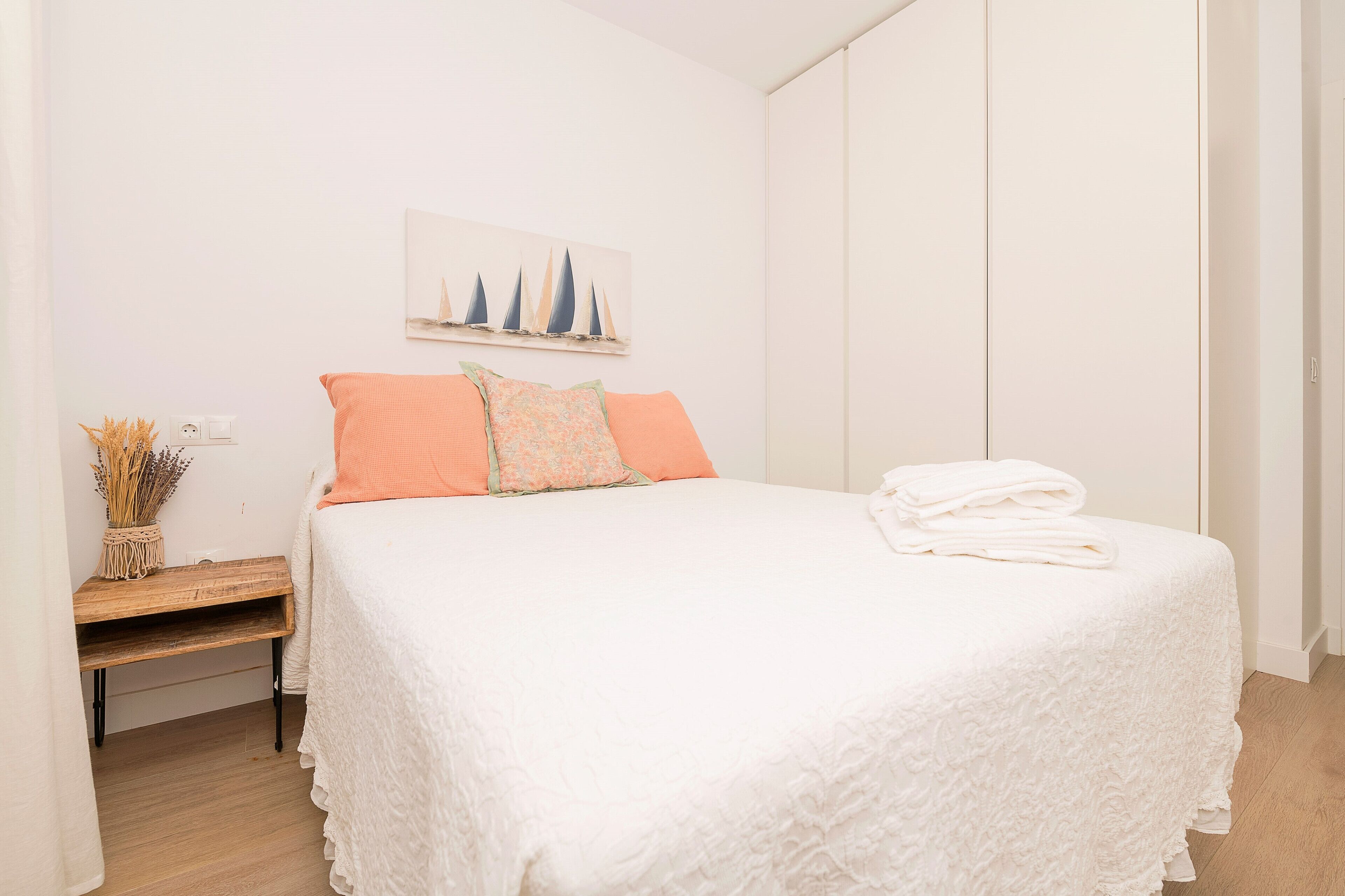 A neatly made bed with a white bedspread and two pillows, a small wooden nightstand with a vase of dried flowers, and a wall-mounted painting of sailboats.