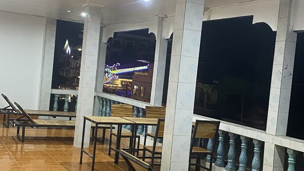 A spacious indoor area with multiple wooden tables and chairs, large windows with decorative balustrades, and a view of a brightly lit building outside.