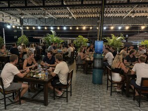 Un área de comedor interior con varias mesas y sillas, rodeada de plantas en macetas y un dispensador de agua.