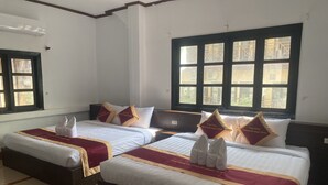 A hotel room with two beds, each with white linens and maroon bedspreads. There are decorative pillows with text, a window with a view of buildings, and a tiled floor.