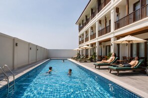 En swimmingpool med liggestole og parasoller, omgivet af en bygning med altaner og vinduer.
