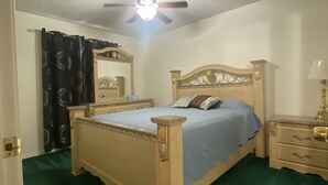 Ein Schlafzimmer mit einem Holzbett, einem Nachttisch, einem Spiegel, einem Deckenventilator und einem grünen Teppich.