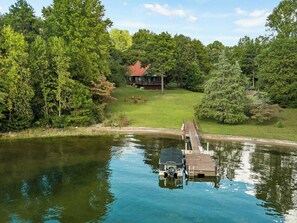 Un lac avec un quai et une maison en arrière-plan, le tout entouré d’arbres.