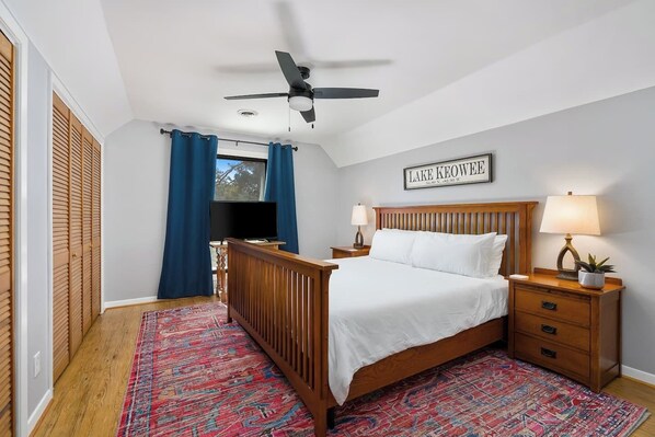 Ein Schlafzimmer mit Bett, Nachttisch, Lampe, Deckenventilator, Fenster mit Vorhängen und einem Schild an der Wand.
