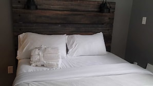 A neatly made bed with white linens and a stack of towels, a wooden headboard, and two wall-mounted lamps.